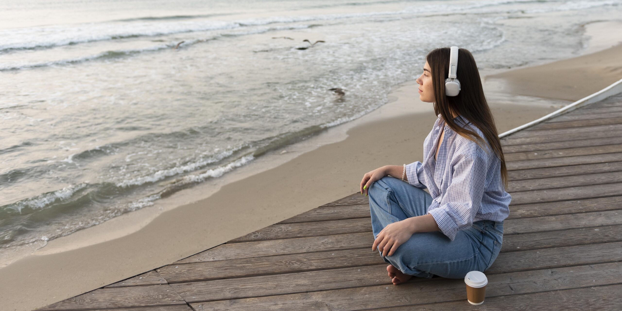 Vista lateral de una mujer joven con audífonos, sentada junto a la playa.